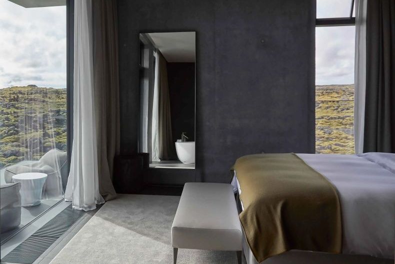 Moss Suite Schlafbereich mit Doppelbett, bodentiefen Fenstern und Blick auf die isländische Lavalandschaft.