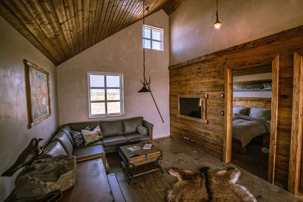 Großzügiger Wohnraum mit Ledersofa, hohem Holzgebälk, Fellteppich und Blick in das Schlafzimmer.