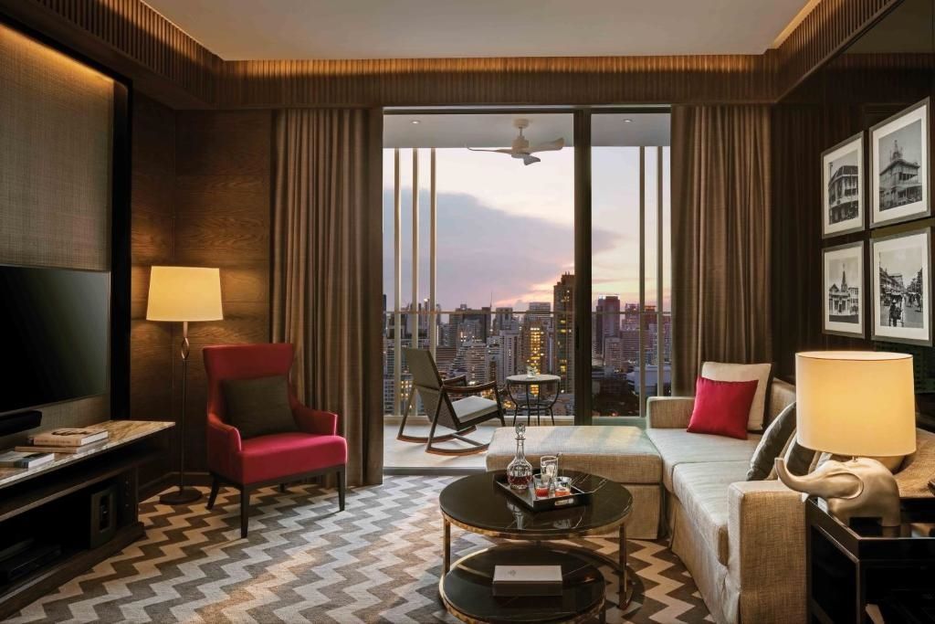 Wohnbereich der Rattanakosin Suite mit stilvollen Möbeln und großem Fenster mit Sonnenuntergang.