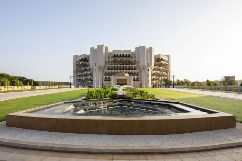 Majestätisches Hauptgebäude des Al Bustan Palace, A Ritz-Carlton Hotel, mit orientalischer Architektur, Palastfassade, gepflegtem Rasen und dekorativem Wasserbecken mit Springbrunnen im Vordergrund.