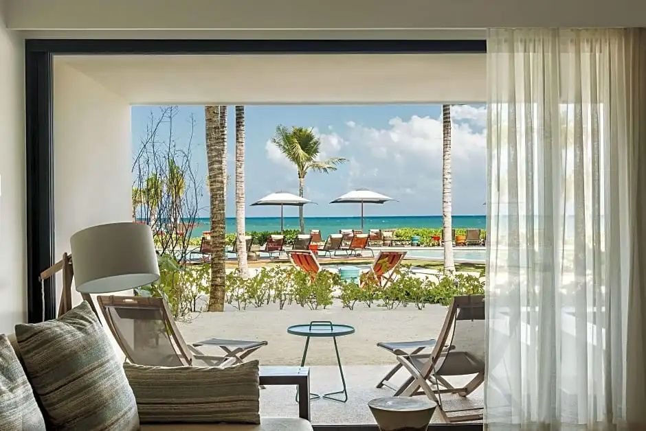 Blick von einer schattigen Terrasse auf den Sandstrand mit Sonnenschirmen, Liegen und Palmen am Meer.