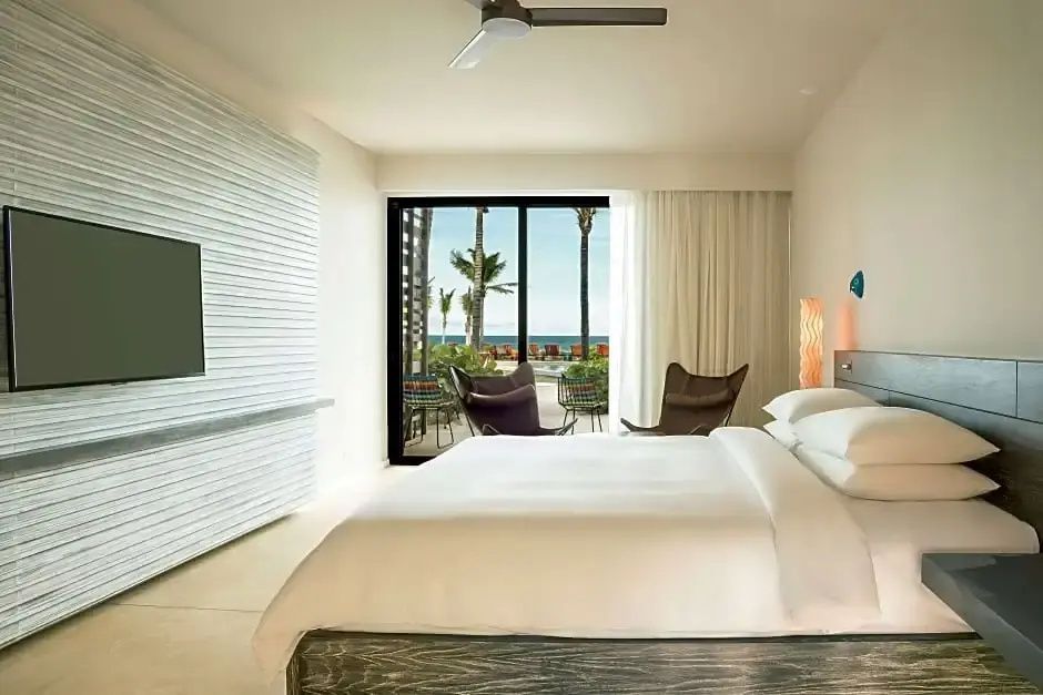 Helles Schlafzimmer mit Doppelbett, Deckenventilator und Blick durch Schiebetüren auf Palmen und das Meer.