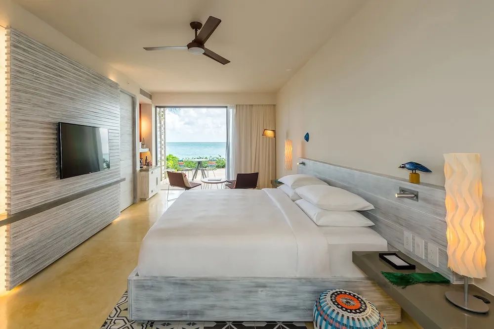 Helles Hotelzimmer mit Doppelbett, Deckenventilator und Zugang zur Terrasse mit Meerblick.
