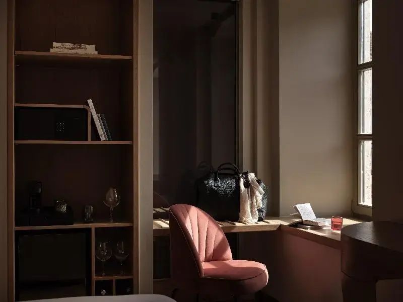 Sitzecke im Hotelzimmer mit rosa Sessel, Holzregal und Arbeitsfläche am Fenster.