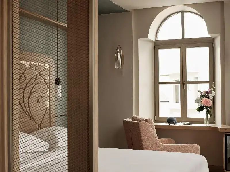 Hotelzimmer mit Doppelbett, Sessel und Rundbogenfenster in hellen Tönen.