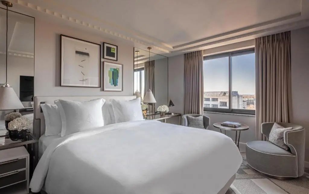 Hell eingerichtetes Hotelzimmer mit Doppelbett, Wandbildern, Sitzbereich und großem Fenster mit Stadtblick.