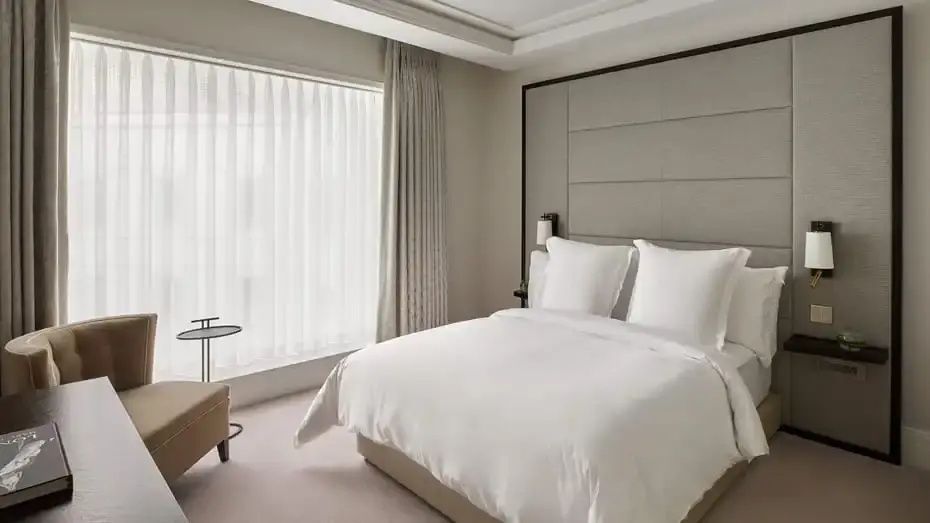 Helles Hotelzimmer mit weiß bezogenem Doppelbett und großem Fenster mit Vorhängen.