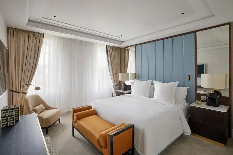 Hotelzimmer mit weiß bezogenem Doppelbett, blauem Kopfteil und orangener Sitzbank.