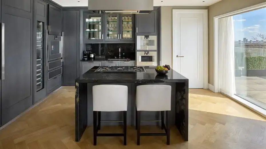 Dunkle Kücheninsel mit Kochfeld und zwei weißen Barhockern in einem modernen Apartment.