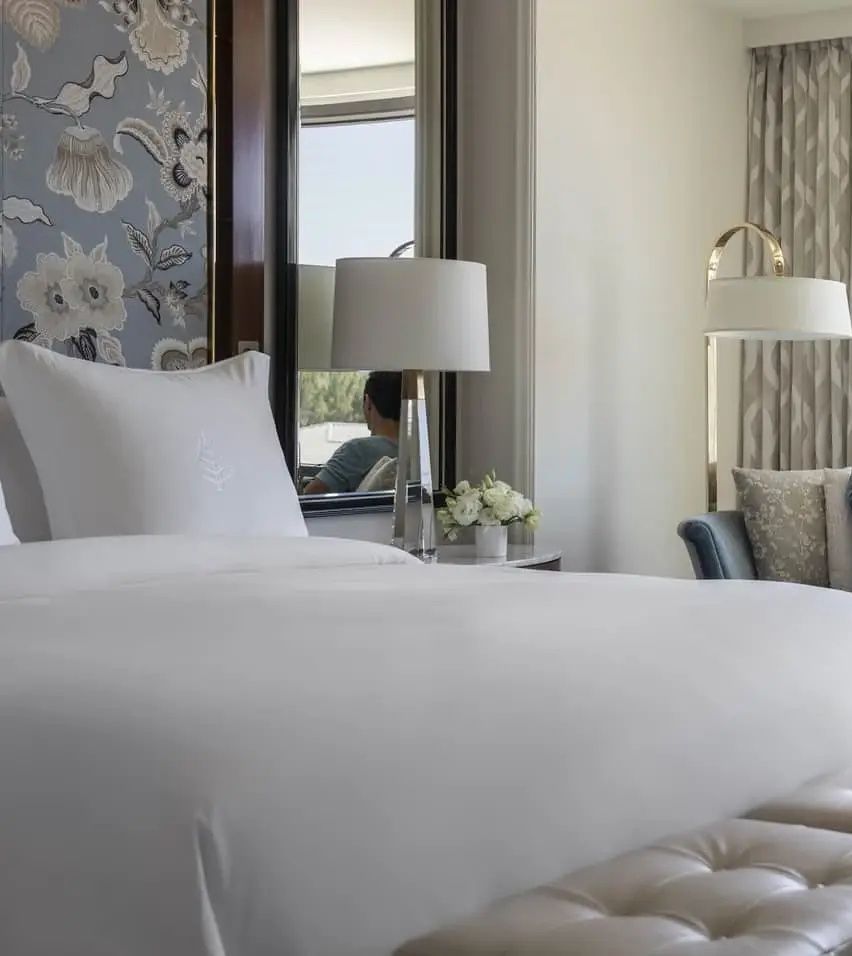 Gemachtes Hotelbett mit weißer Bettwäsche, Nachttischlampe und floral gemusterter Tapete.