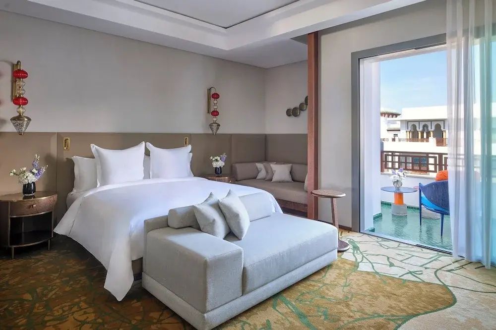 Helles Hotelzimmer mit weißem Doppelbett, gepolsterter Bank, Sitzecke und Balkon.