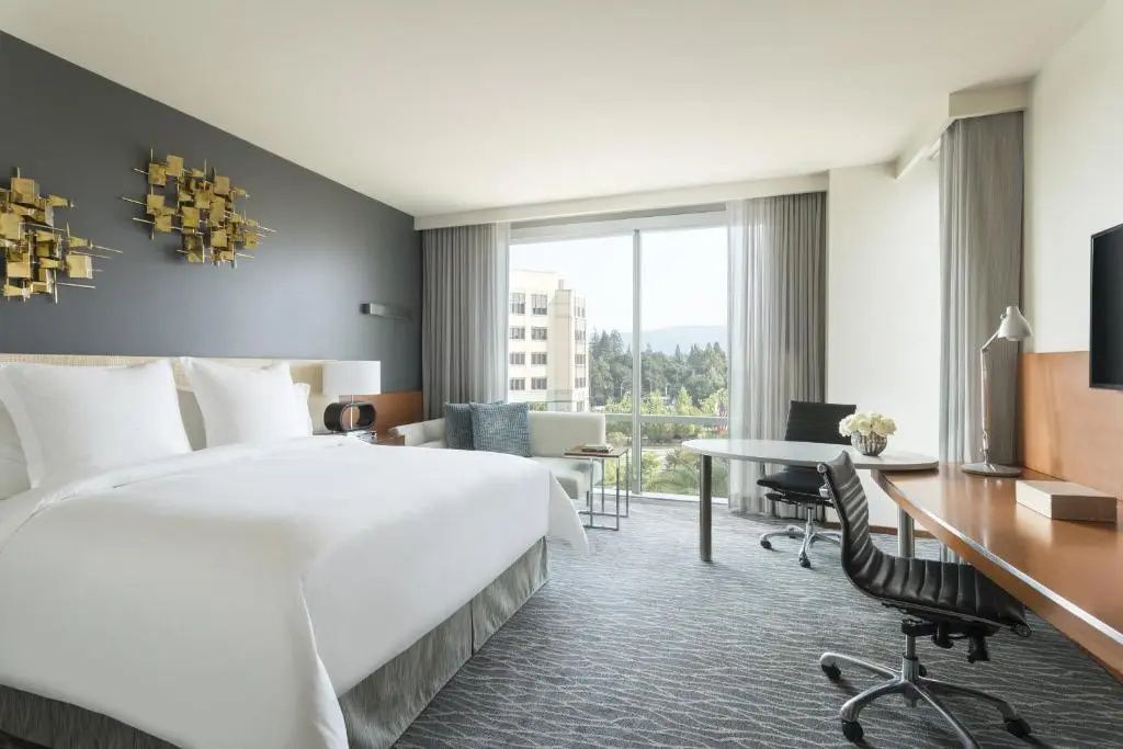 Helles Hotelzimmer mit weißem Bett, Schreibtisch und Fensterfront mit Blick auf Bäume.