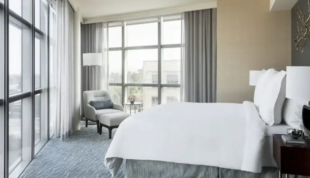Helles Hotelzimmer mit weiß bezogenem Bett, einem grauen Sessel und großen Fensterfronten.
