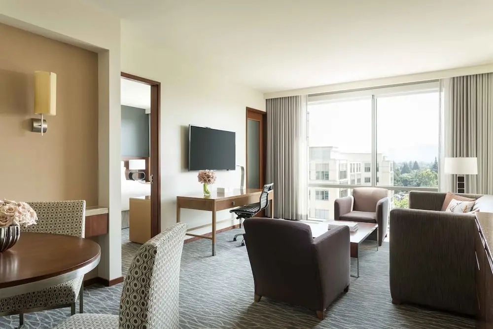 Wohnbereich einer Suite mit Holzschreibtisch, Wandfernseher und zwei Sesseln am Fenster.