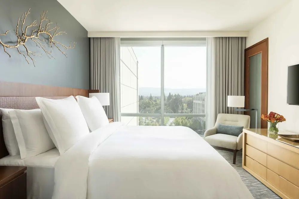 Hotelzimmer mit weiß bezogenem Doppelbett, Sessel und Blick durch das Fenster ins Grüne.