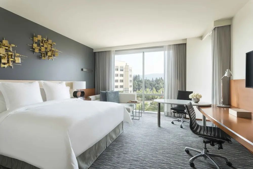 Hotelzimmer mit großem Bett, Schreibtisch am Fenster und Blick auf Gebäude im Hintergrund.