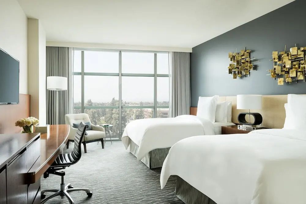 Hotelzimmer mit zwei weißen Betten, Schreibtisch und großer Fensterfront.