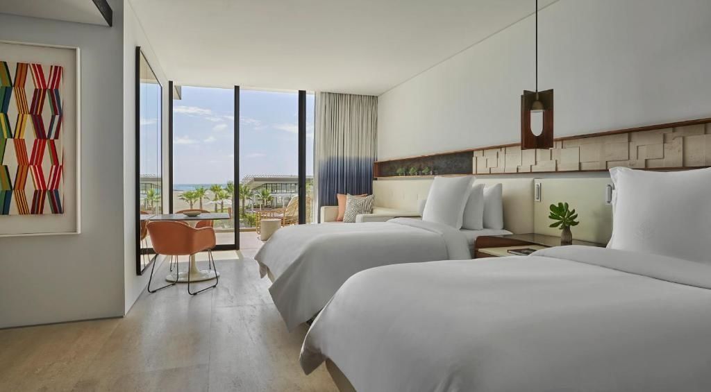 Helles Hotelzimmer mit zwei Doppelbetten, Sitzbereich und großer Glasfront mit Blick auf das Meer.