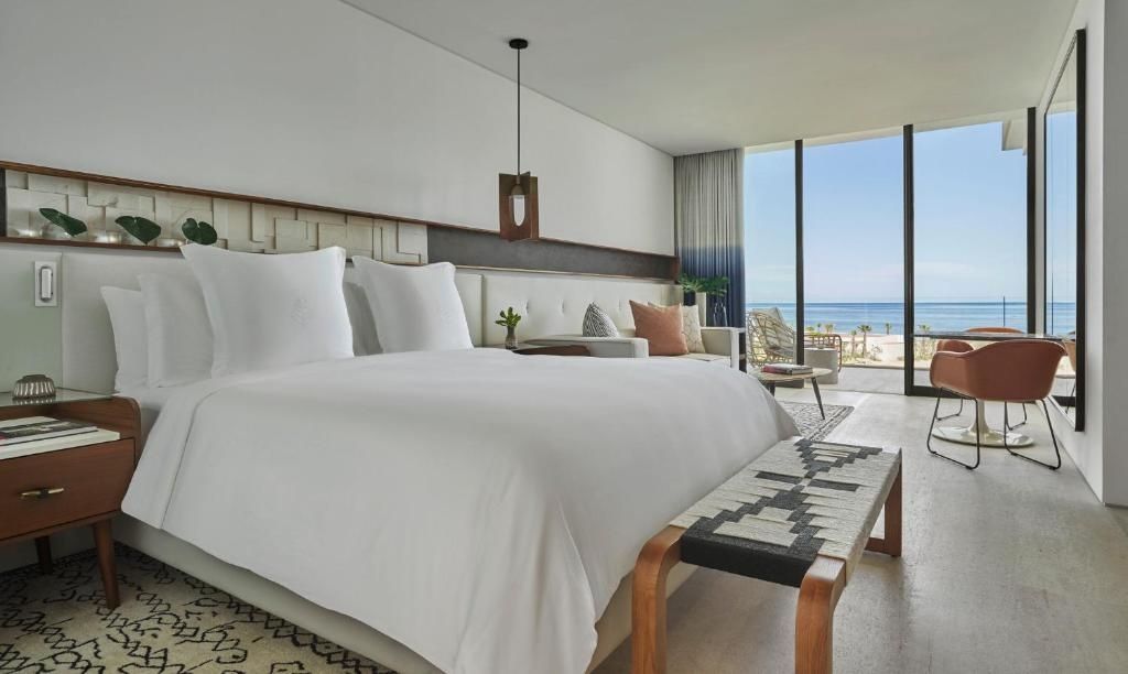 Modernes Schlafzimmer mit weißem Doppelbett, Holzbank und direktem Blick auf das Meer.