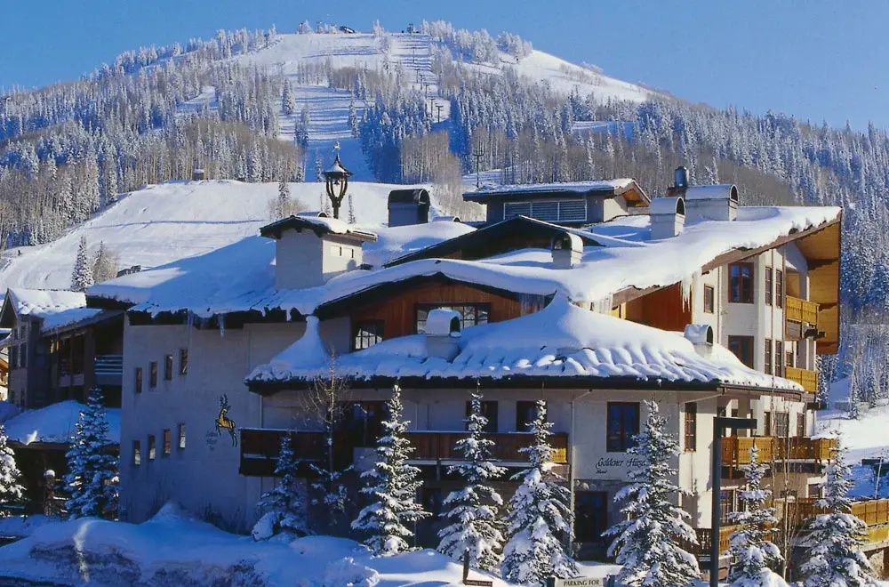 Verschneites Hotelgebäude im alpenländischen Stil vor verschneiten Bergen und Skipisten.