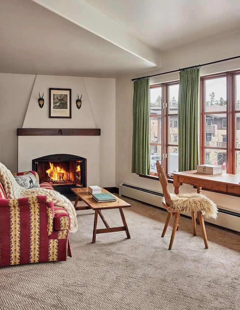 Wohnzimmer mit brennendem Kamin, gemustertem Sofa, Holztisch und Schreibtisch vor großem Fenster.