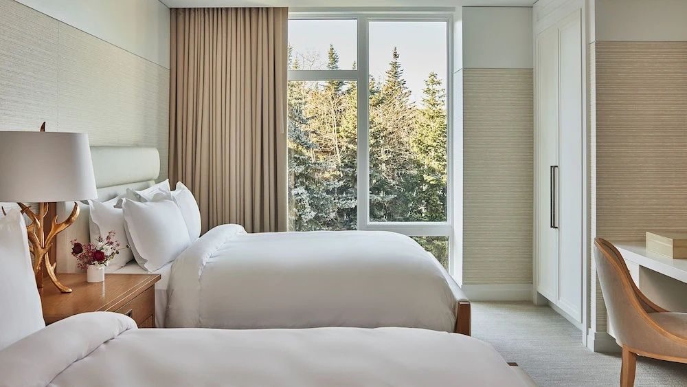 Hotelzimmer mit weiß bezogenen Betten und einem großen Fenster mit Blick auf Nadelbäume.