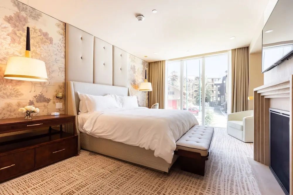 Helles Hotelzimmer mit Doppelbett, beige gepolstertem Kopfteil und großer Fensterfront.