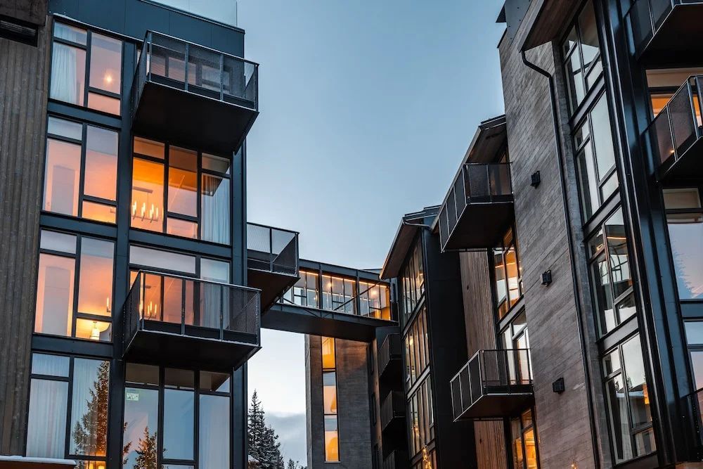 Moderne Holz- und Metallfassade des Gebäudes mit beleuchteten Fenstern und Balkonen bei Dämmerung.