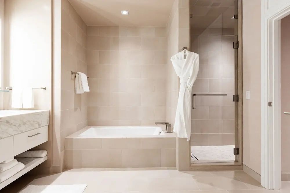 Hell gefliestes Badezimmer mit einer Badewanne, gläserner Duschkabine und einem Bademantel.