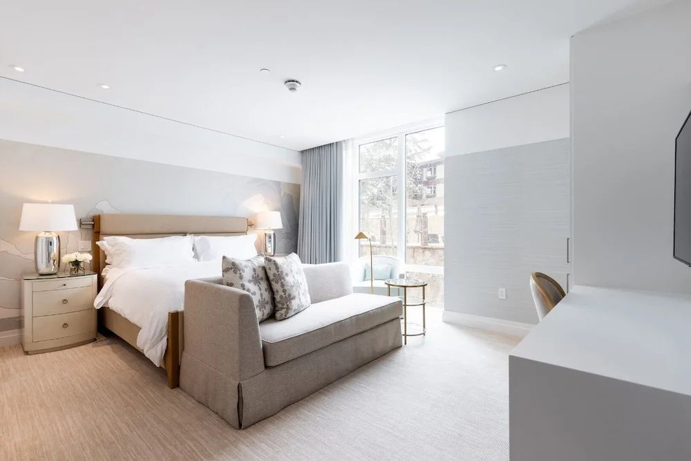 Helles Hotelzimmer mit großem Doppelbett, grauem Sofa am Bettende und Fensterfront.