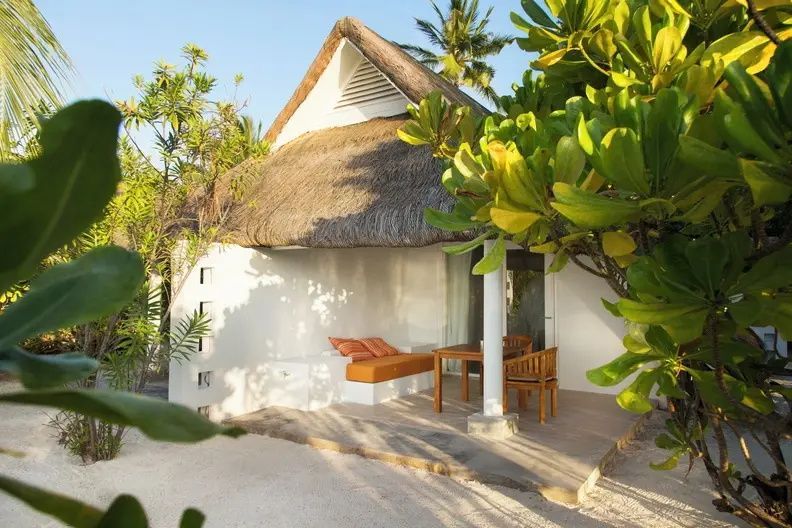 Strandvilla mit Reetdach, überdachter Terrasse, Holzmöbeln und Tropenpflanzen an einem Sandstrand.