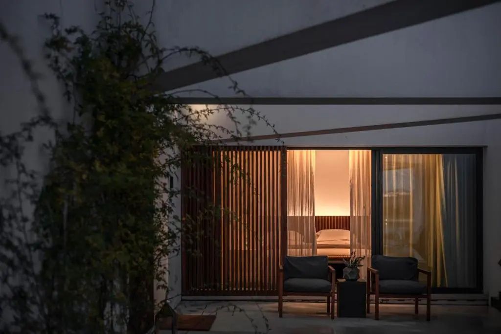 Überdachte Terrasse mit zwei Sesseln und Blick durch ein Fenster in ein beleuchtetes Schlafzimmer.