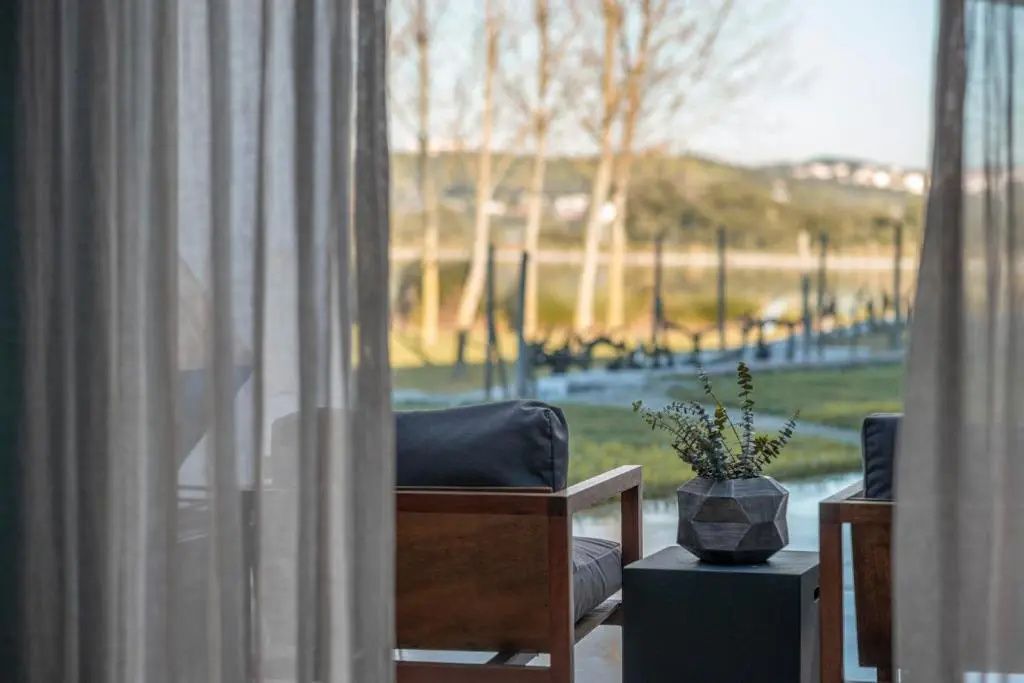 Blick durch transparente Vorhänge auf einen Sessel, eine Vase und die Landschaft mit See.