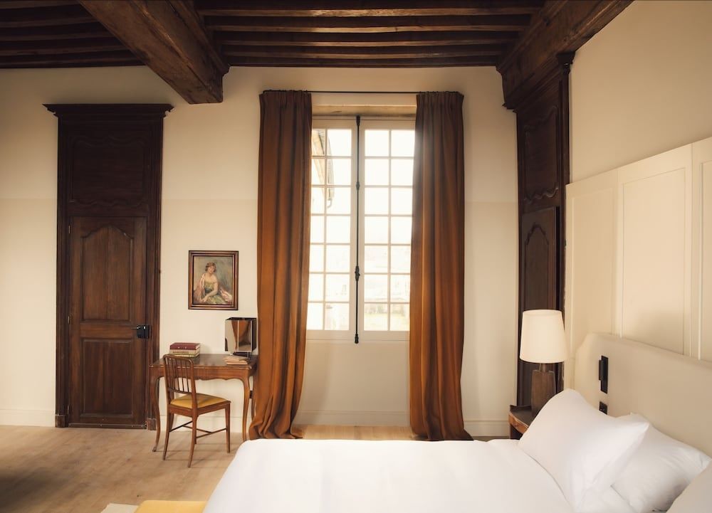 Hotelzimmer mit hohem Fenster, braunen Vorhängen, Holzbalken und Schreibtisch.