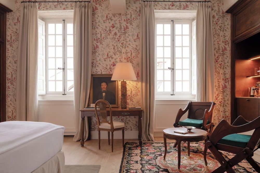 Hotelzimmer mit blumiger Tapete, zwei Fenstern, einem Schreibtisch und Sesseln.