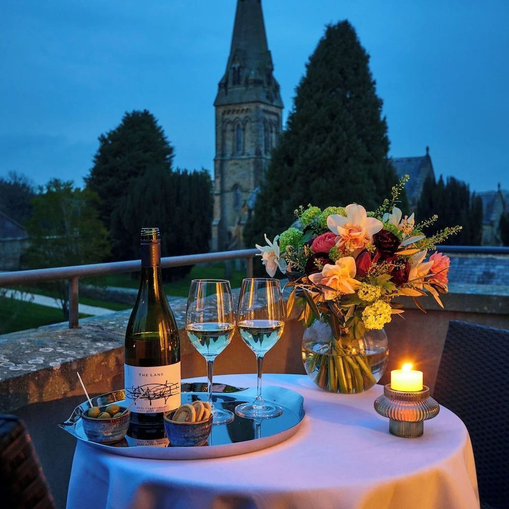 Gedeckter Tisch auf einer Terrasse mit Wein, Blumen und Blick auf einen Kirchturm am Abend.