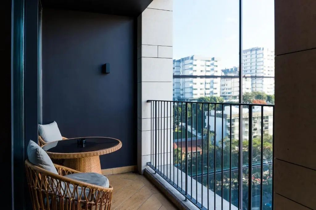 Balkon mit Rattansesseln und Tisch mit Blick auf Stadtgebäude und grüne Bäume.