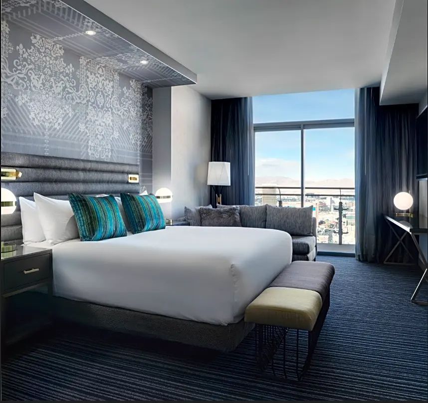 Helles Hotelzimmer mit großem Doppelbett, grauem Sofa und Balkontür mit Blick auf die Stadt.