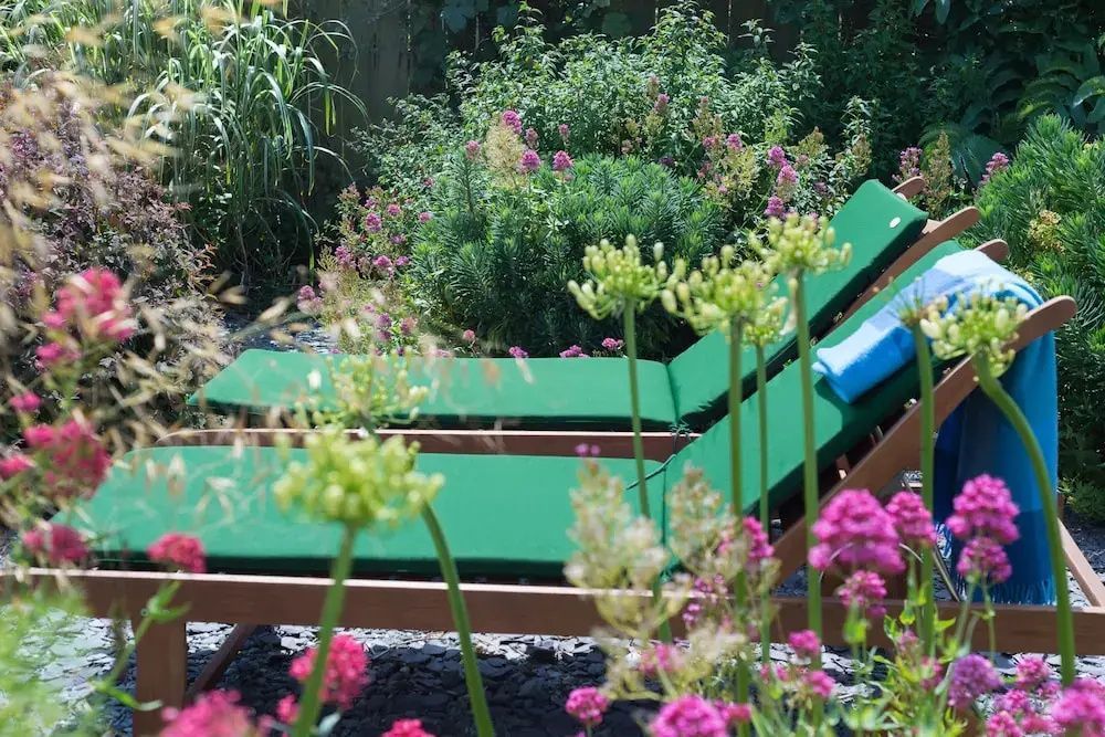 Zwei hölzerne Sonnenliegen mit grünen Polstern inmitten von blühenden Gartenpflanzen.