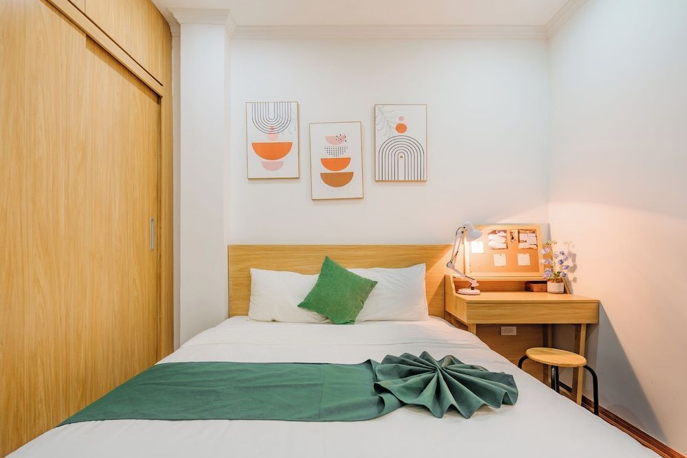 Doppelbett mit grüner Tagesdecke, Holzkopfteil und Schreibtisch im hellen Zimmer.