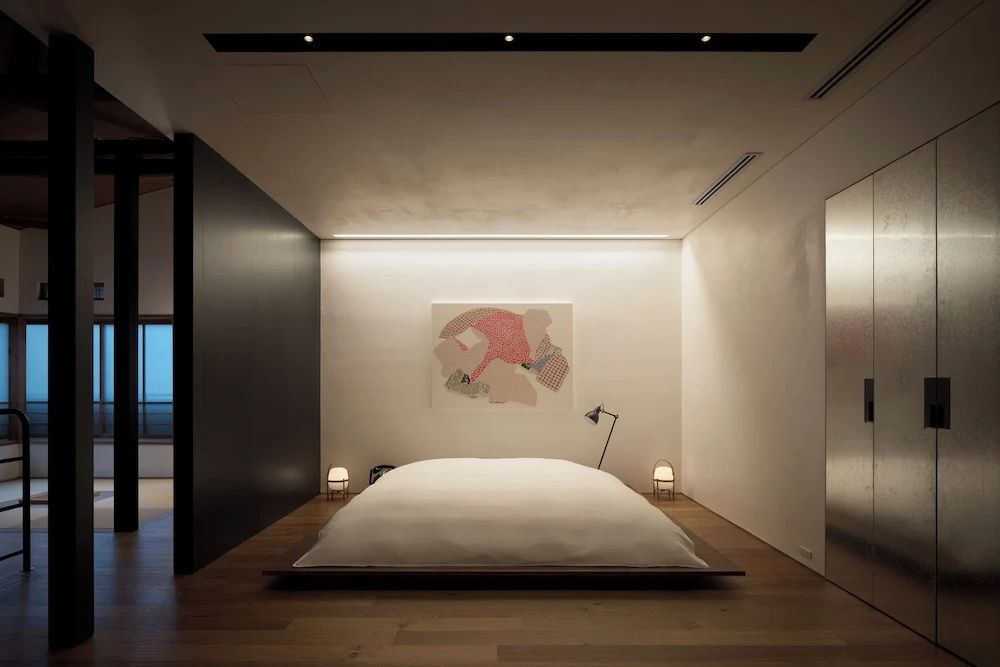 Schlafbereich mit bodennahem Bett, heller Wandgestaltung, Kunstwerk und dezenter Beleuchtung.