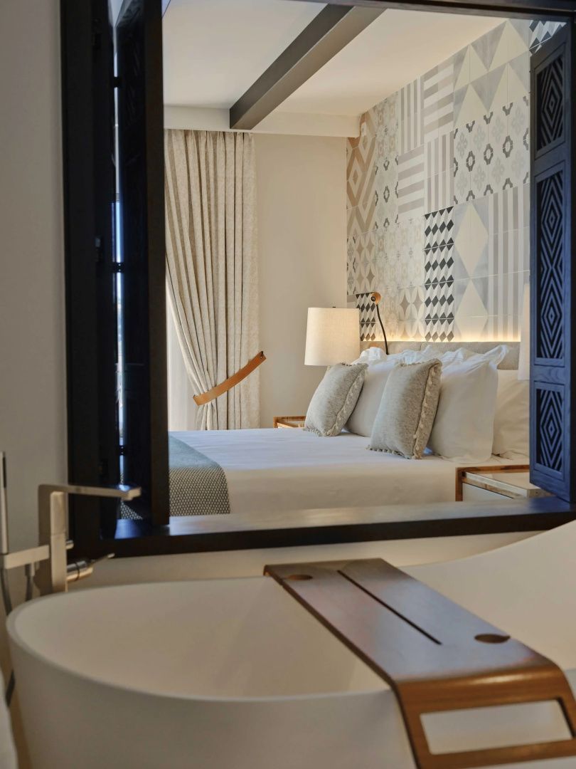 Blick durch ein Wandfenster auf ein Doppelbett mit hellen Kissen und gemusterter Wand im Zimmer.