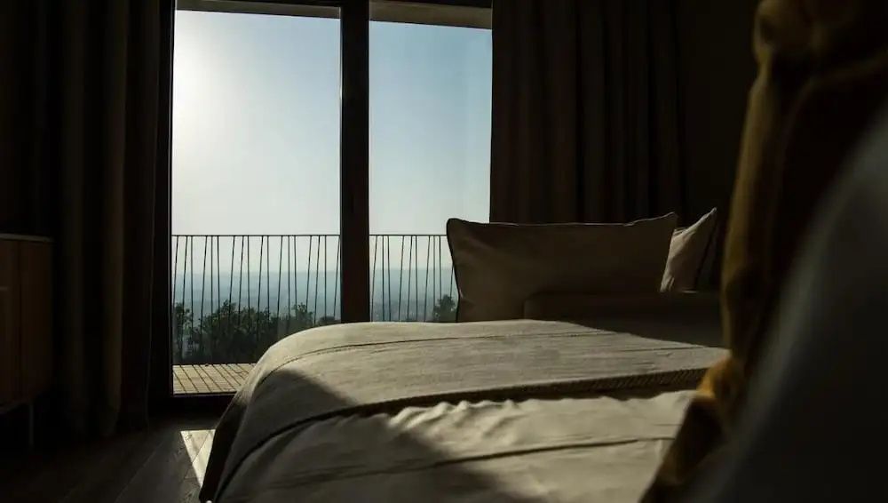 Blick aus dem Hotelzimmer mit gemachtem Bett durch ein großes Fenster auf den Balkon und die Landschaft.