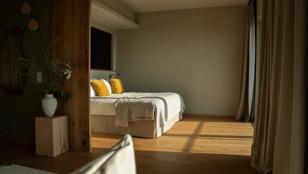 Doppelbett mit weißer Bettwäsche und gelben Kissen in einem sonnigen Zimmer mit Holzboden.