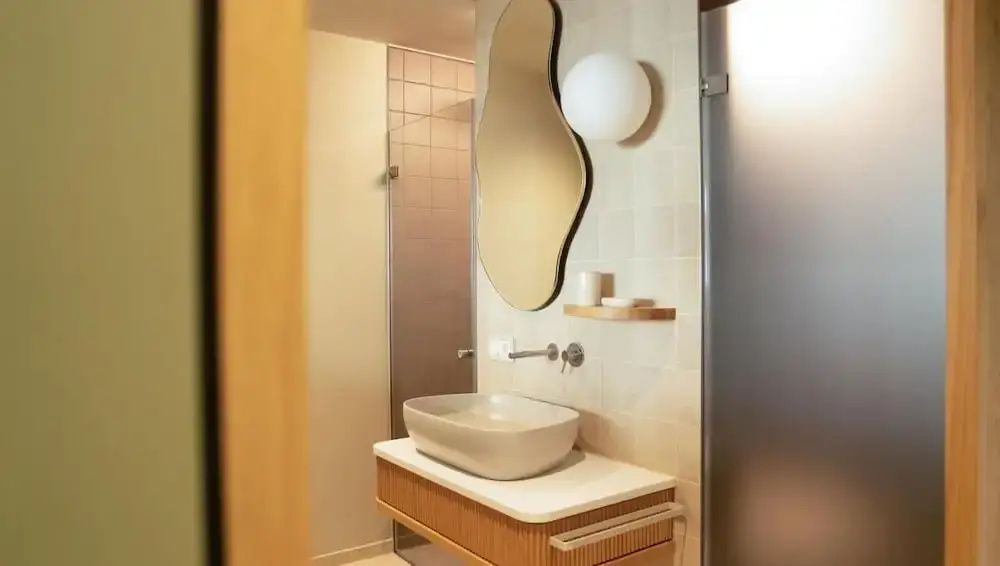 Badezimmer mit einem ovalen Waschbecken auf einem Holzschrank und einem organisch geformten Spiegel.