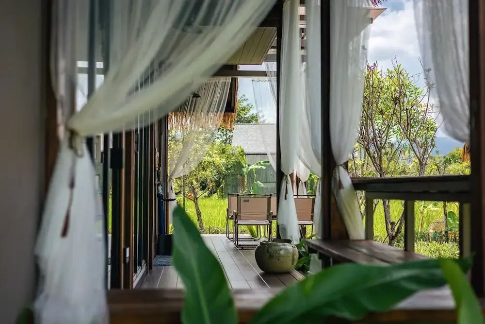Überdachte Holzterrasse mit weißen Vorhängen und Blick ins Grüne auf Gartenstühle.