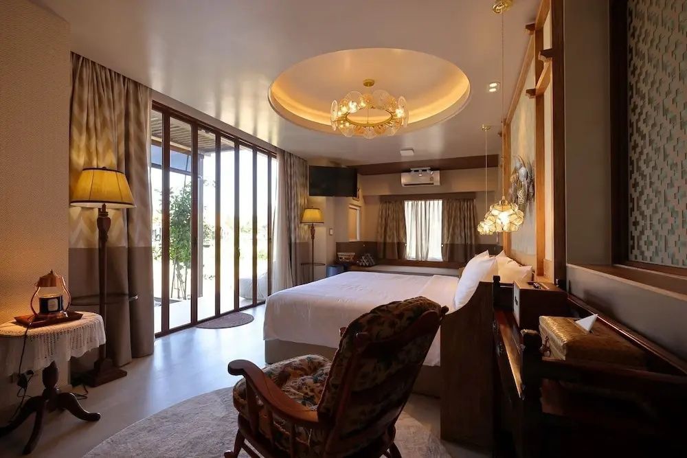Geräumiges Hotelzimmer mit großem Doppelbett, Lehnstuhl und bodentiefen Glasfenstern.