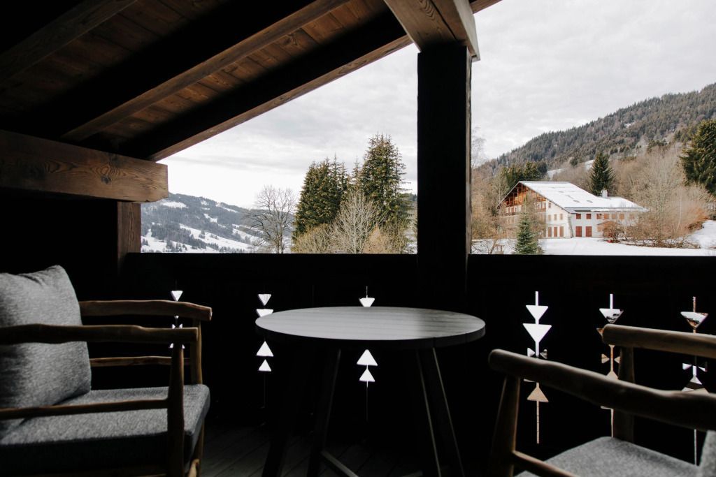 Balkon mit Blick auf verschneite Berge und Wälder. Schwarzer Tisch und Stühle im Vordergrund.