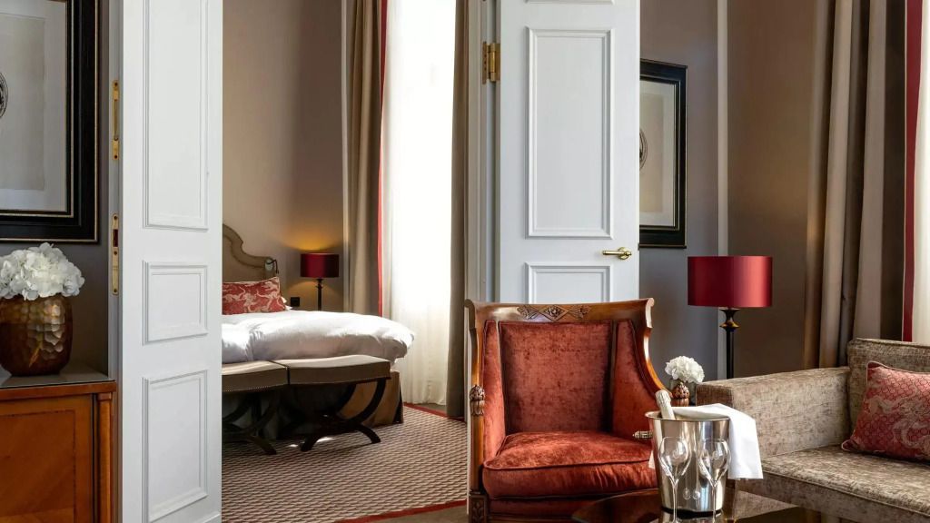 Elegante Suite mit gemütlichem Sessel, Champagnerkühler und Doppelbett im Hintergrund.