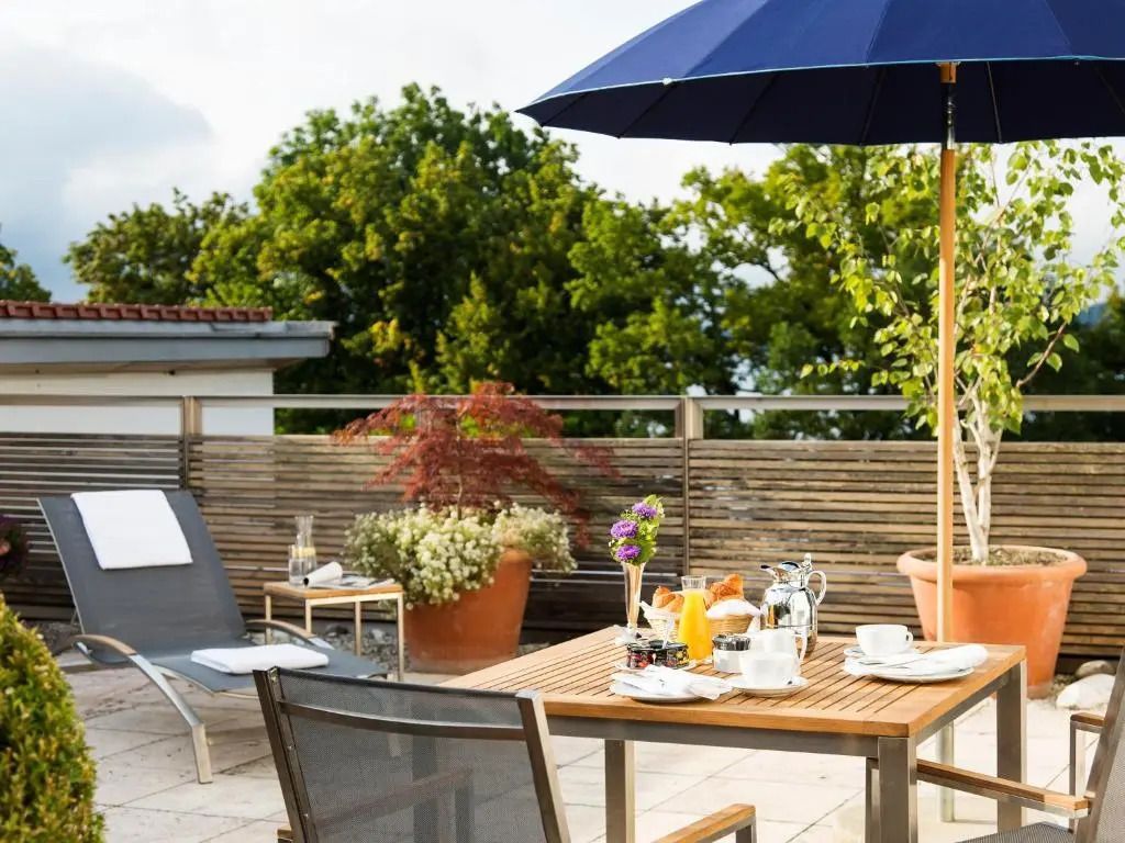 Terrasse mit Frühstückstisch und Sonnenschirm, umgeben von grünen Pflanzen.
